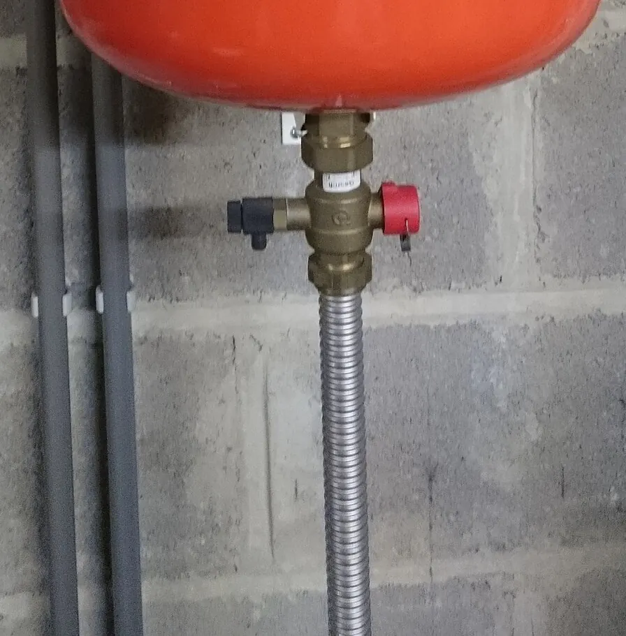 Brass shutoff valve with a small drain valve below a red expansion tank on corrugated pipe
