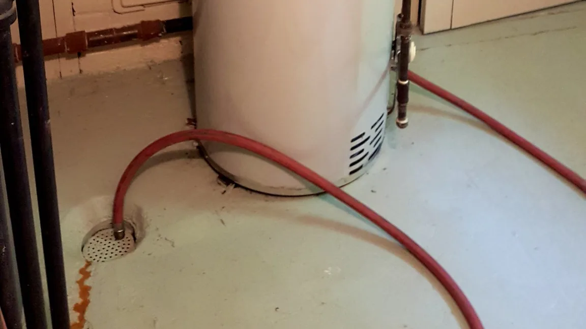 Base of a tank water heater with a red garden hose running from its drain valve to a floor drain