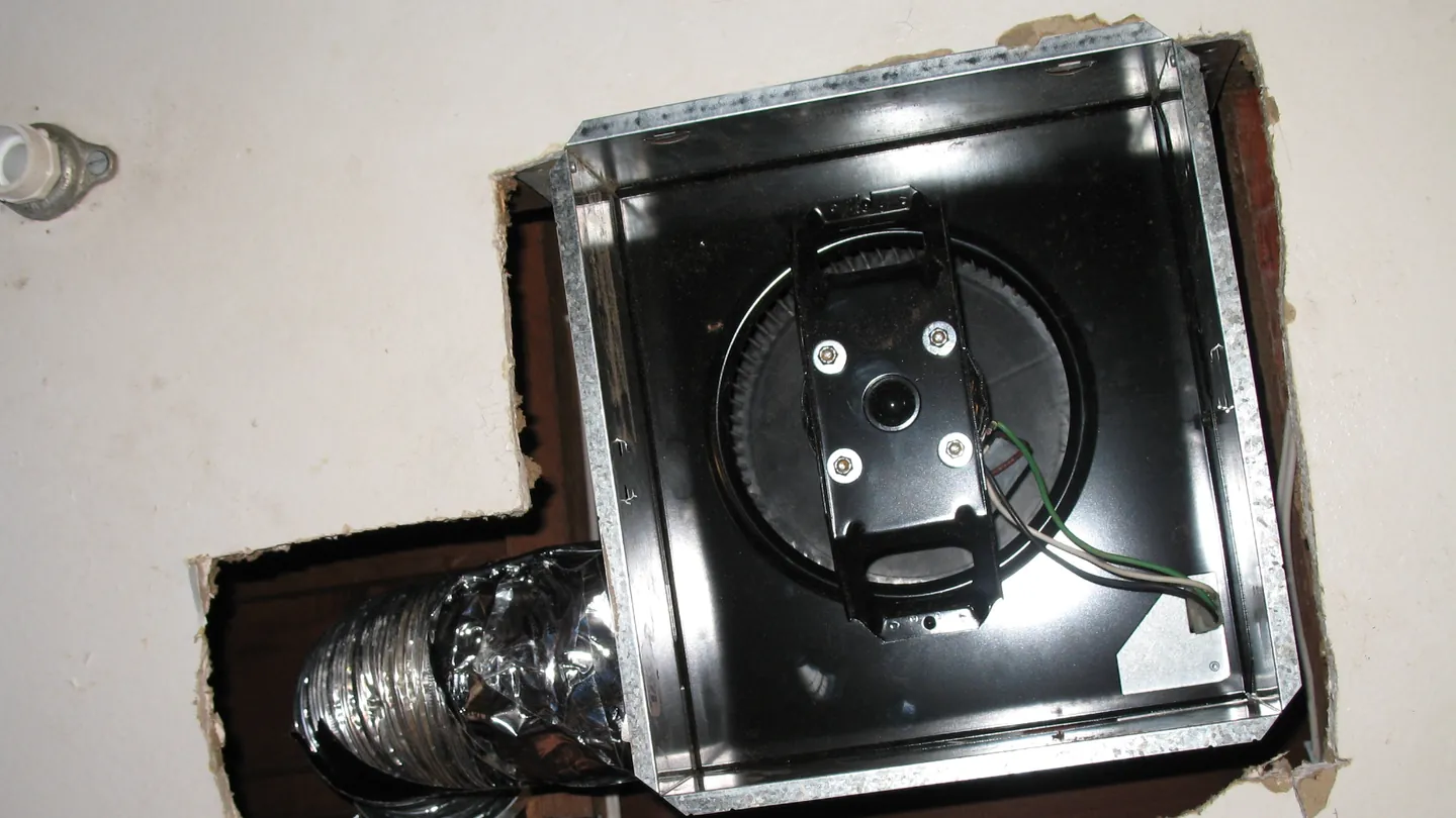 Bathroom exhaust fan housing set into an opening in the ceiling drywall, with its blower and flex duct in place
